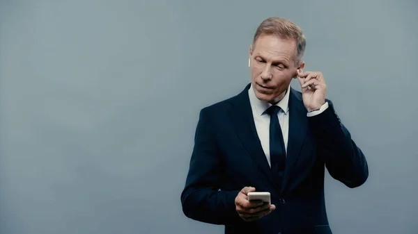 Mature businessman in formal wear using smartphone and earphones isolated on grey — Stock Photo