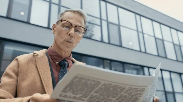 Businessman in coat reading newspaper near blurred building outdoors — Fotografia de Stock