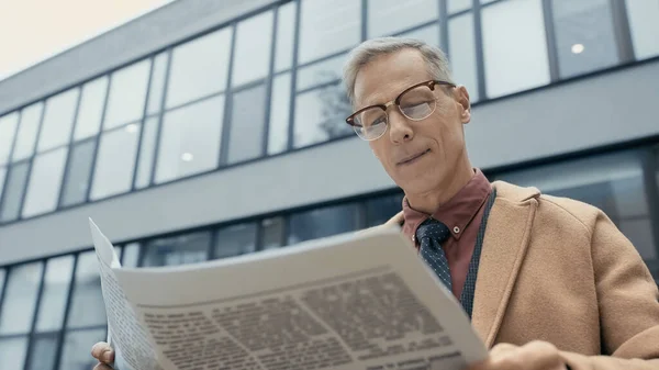 Vista de ángulo bajo del hombre de negocios sonriente en abrigo leyendo el periódico al aire libre - foto de stock