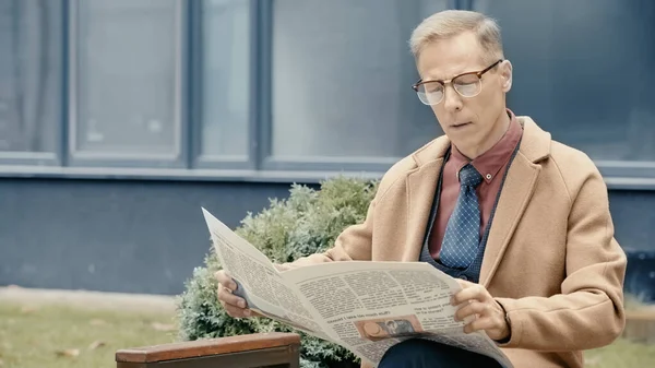 Businessman in coat reading newspaper on urban street — Stock Photo