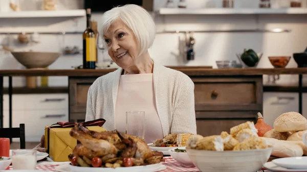 Mujer mayor sonriendo cerca de la caja de regalo y cena de acción de gracias servida en la mesa en la cocina - foto de stock