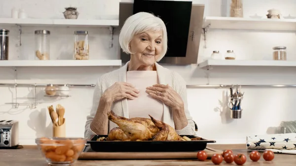 Радісна жінка дивиться на смачну індичку біля свіжих вишневих помідорів і моркви на кухні — стокове фото