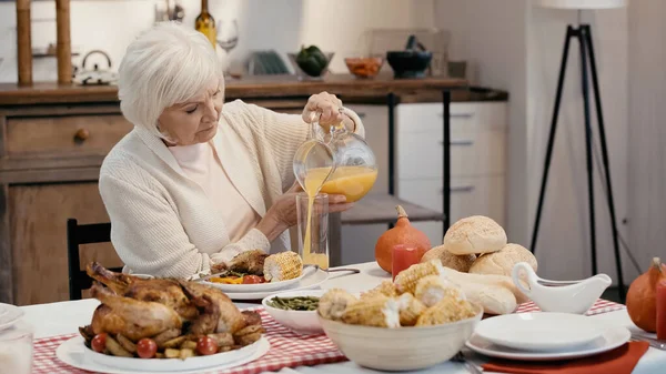 Senior hombre verter jugo de naranja cerca de pavo asado, maíz a la parrilla y bollos en la mesa - foto de stock