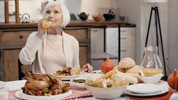 Mujer mayor bebiendo jugo de naranja cerca de pavo asado, maíz a la parrilla y bollos en la mesa - foto de stock