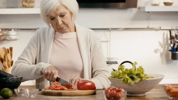 Mujer mayor corte fresco pimiento cerca de lechuga y tomates cherry en la cocina - foto de stock