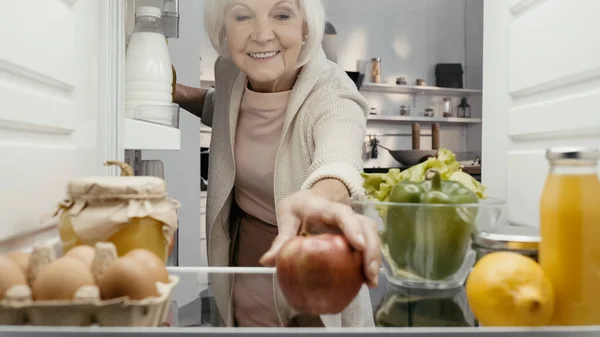 Happy senior woman taking fresh apple from fridge with fresh vegetables, fruits, drinks and eggs — Stock Photo