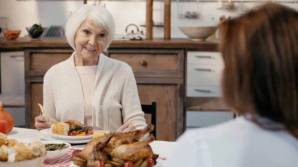 Senior woman smiling near blurred guest during thanksgiving dinner in kitchen — стоковое фото