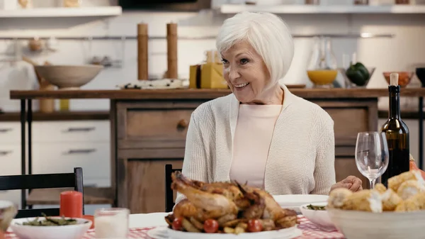 Happy senior woman sitting near roasted turkey and red wine during thanksgiving dinner — Foto stock