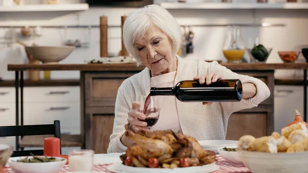 Elderly woman pouring red wine near thanksgiving dinner served in kitchen — стоковое фото