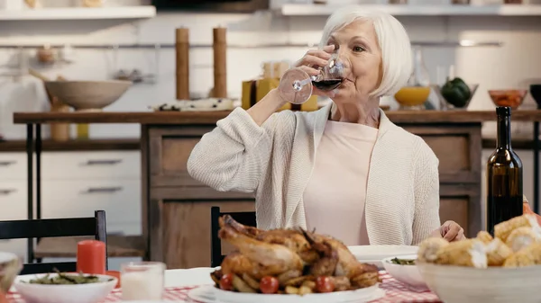 Mujer mayor con pelo gris bebiendo vino tinto cerca de pavo asado durante la cena de Acción de Gracias - foto de stock