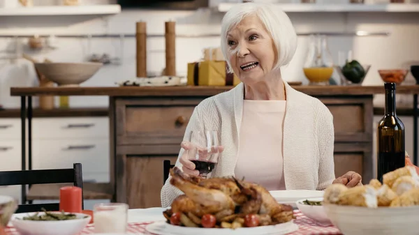 Happy senior woman holding glass of red wine near traditional roasted turkey during thanksgiving dinner — Fotografia de Stock