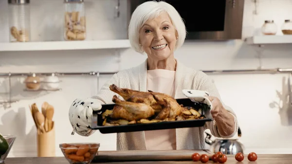 Happy senior woman holding oven sheet with roasted turkey near fresh cherry tomatoes and carrots in kitchen — Foto stock