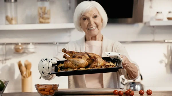 Happy senior woman holding oven sheet with delicious turkey and potatoes near fresh vegetables, blurred background — Foto stock