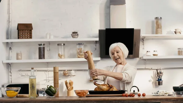 Joyeuse femme âgée assaisonnement dinde rôtie près des légumes frais et de l'huile dans la cuisine — Photo de stock