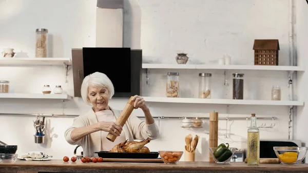 Smiling senior woman seasoning roasted turkey near fresh vegetables and oil on table — стоковое фото