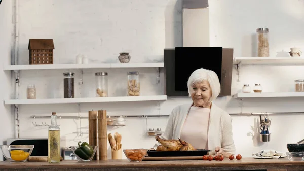 Pleased senior woman looking at delicious turkey near fresh vegetables and spice mills in kitchen — стоковое фото