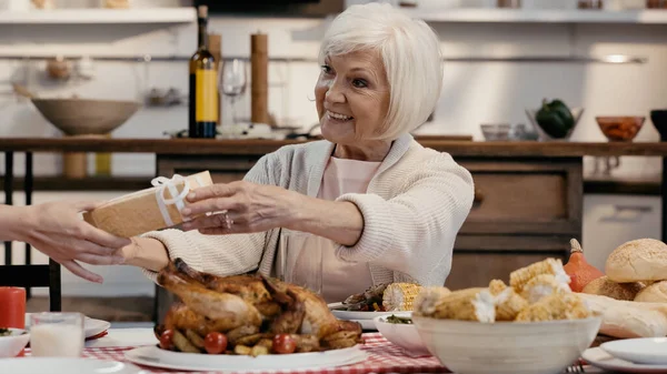 Invitado dando regalo a sonriente mujer mayor sentada en la mesa servida con cena de acción de gracias - foto de stock
