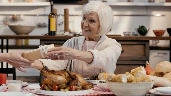 Guest giving present to smiling senior woman sitting at table served with thanksgiving dinner — Fotografia de Stock