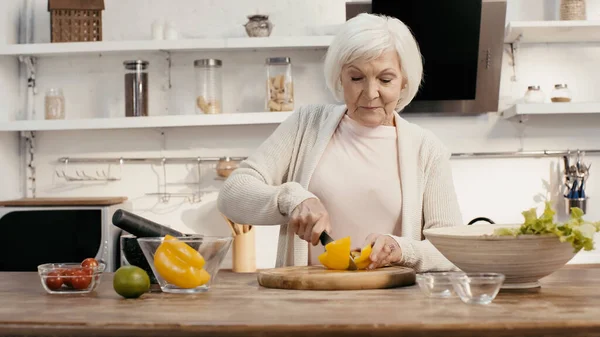 Mujer mayor corte pimiento cerca de cuencos con verduras frescas en la cocina - foto de stock