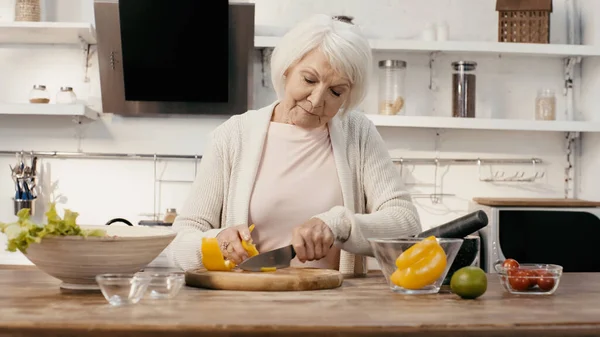 Senior woman cutting bell pepper while preparing vegetable salad in kitchen — Foto stock