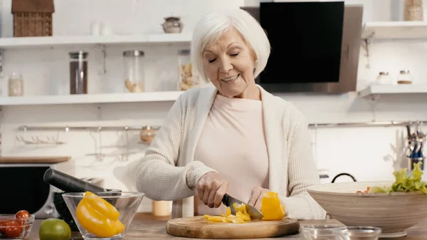Elderly woman smiling while cutting fresh bell pepper on chopping board — стоковое фото