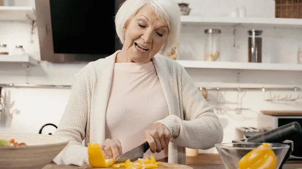 Cheerful senior woman cutting fresh bell pepper on chopping board in kitchen — Foto stock