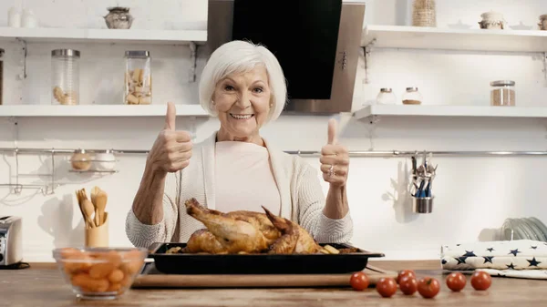 Mujer feliz mostrando pulgares hacia arriba cerca de pavo asado y verduras frescas en la mesa de la cocina - foto de stock