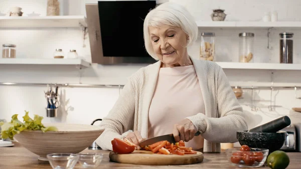 Alegre mujer mayor corte pimiento cerca de tomates cherry frescos y lima en la cocina - foto de stock