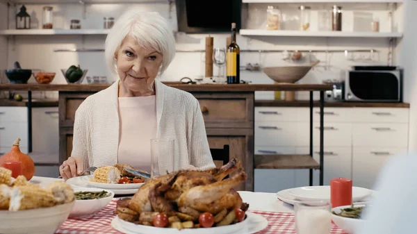 Mujer mayor positiva sentada en la mesa servida con cena de acción de gracias cerca de huésped borrosa - foto de stock
