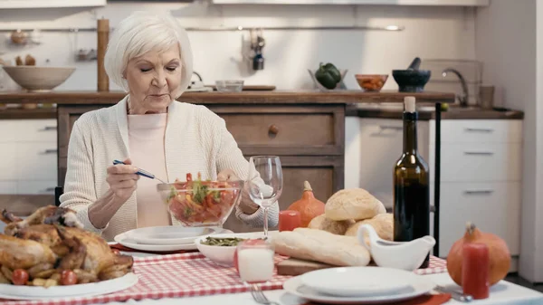 Старша жінка тримає миску овочевого салату біля смаженої індички та червоного вина на столі — стокове фото