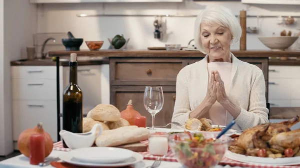 Smiling senior woman praying before thanksgiving dinner near roasted turkey with vegetables, red wine and buns — Fotografia de Stock