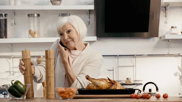 Positive senior woman talking on mobile phone near roasted turkey, spice mills and fresh vegetables — стоковое фото