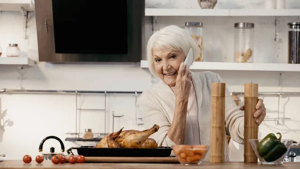 Cheerful senior woman talking on cellphone near roasted turkey, spice mills and fresh vegetables — Stock Photo