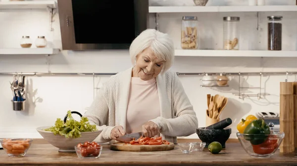 Feliz mujer mayor corte pimiento cerca de tomates cherry frescos, lechuga y zanahorias - foto de stock