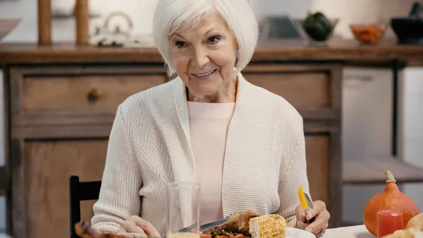 Senior woman smiling near roasted meat and grilled corn during thanksgiving dinner — стоковое фото
