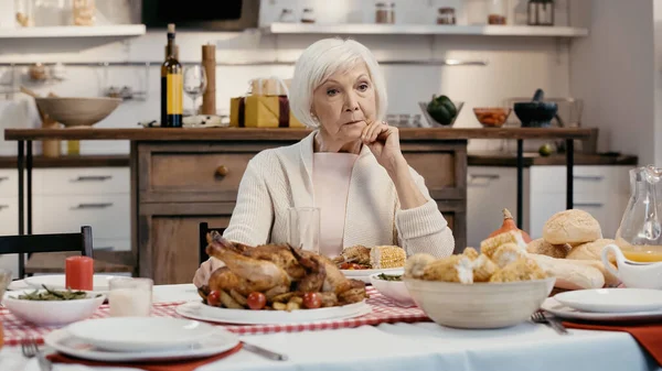 Mujer anciana solitaria y pensativa sentada cerca de la cena de acción de gracias servida en la mesa en la cocina - foto de stock