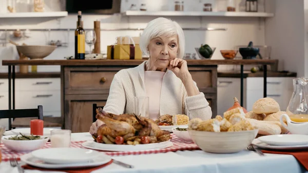 Upset senior woman sitting alone near thanksgiving dinner on table in kitchen - foto de stock
