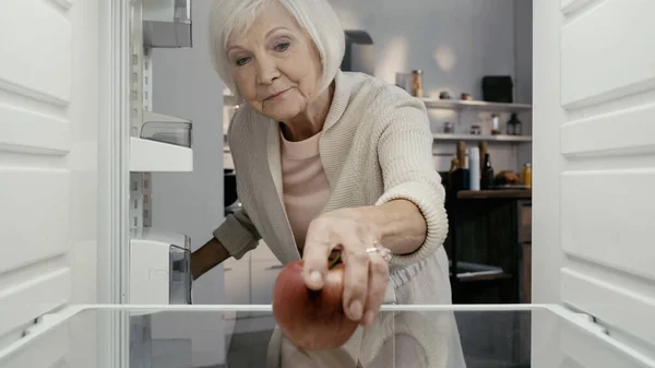 Старша жінка бере стигле червоне яблуко з холодильника на кухні — стокове фото