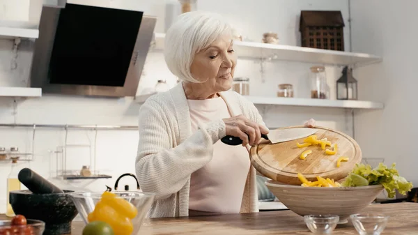 Positive senior woman preparing vegetable salad and adding sliced bell pepper into bowl — Foto stock