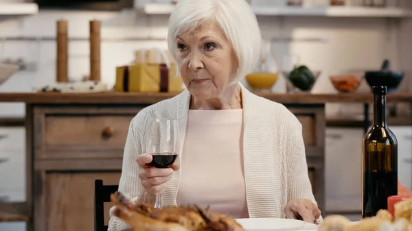Senior woman holding glass of red wine near blurred roasted turkey during thanksgiving dinner — стоковое фото