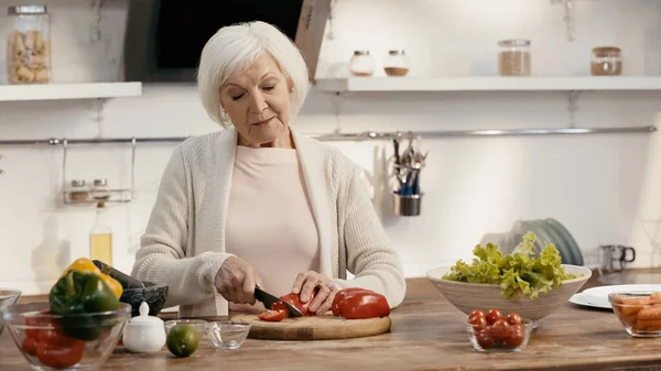 Старша жінка, що ріже болгарський перець біля свіжих овочів на кухонному столі — стокове фото
