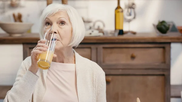 Elderly woman with grey hair drinking orange juice in blurred kitchen — Foto stock