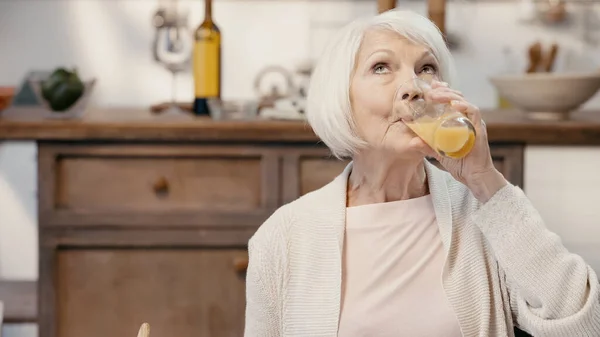 Senior woman drinking orange juice in blurred kitchen — Fotografia de Stock