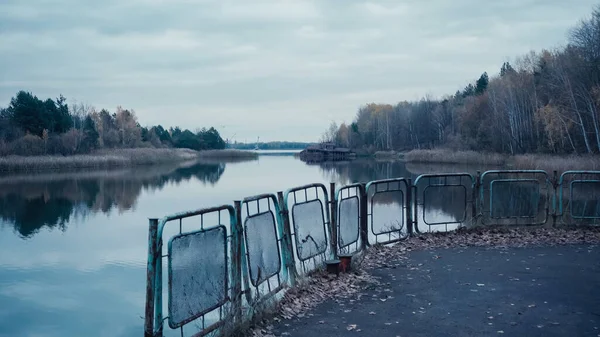 Pripyat river and embankment with rusty fence under grey cloudy sky in chernobyl exclusion zone — стоковое фото