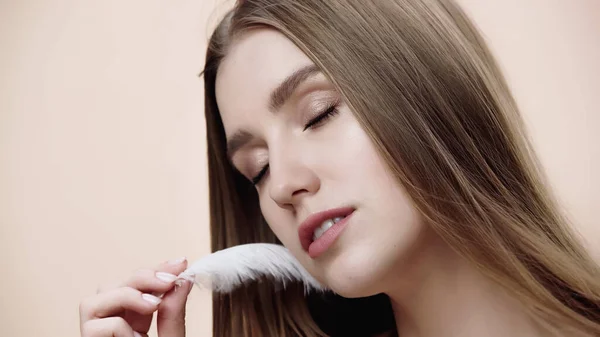 Sensual young woman with closed eyes holding feather near face isolated on beige — стоковое фото