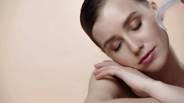 Soft feather near cheek of pretty young woman with closed eyes isolated on beige — Stock Photo