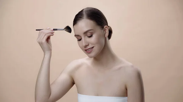 Pleased young woman with bare shoulders applying face powder while holding cosmetic brush isolated on beige — Photo de stock