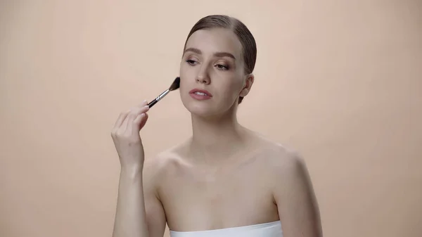 Woman with bare shoulders applying face powder while holding cosmetic brush isolated on beige — стоковое фото