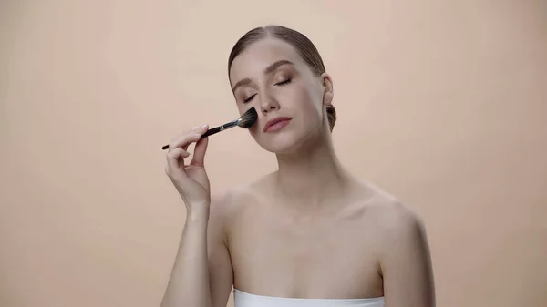 Young woman with bare shoulders and closed eyes applying face powder isolated on beige — стоковое фото