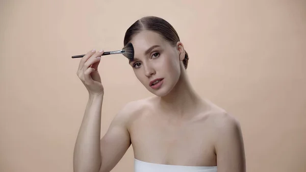 Young woman applying face powder while holding cosmetic brush isolated on beige — Fotografia de Stock